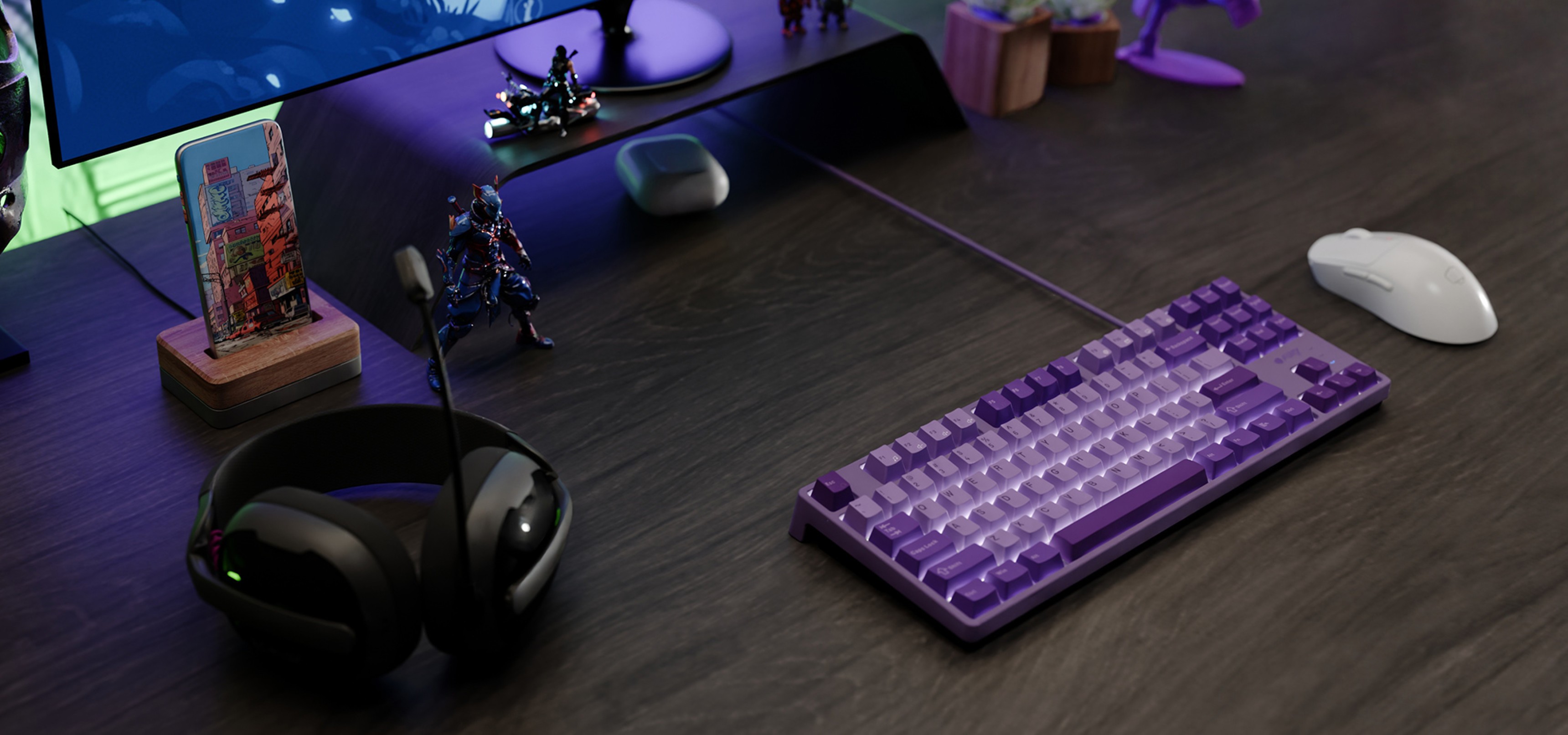 A purple keyboard and a white mouse on a dark wood desk with headphones and figurines.